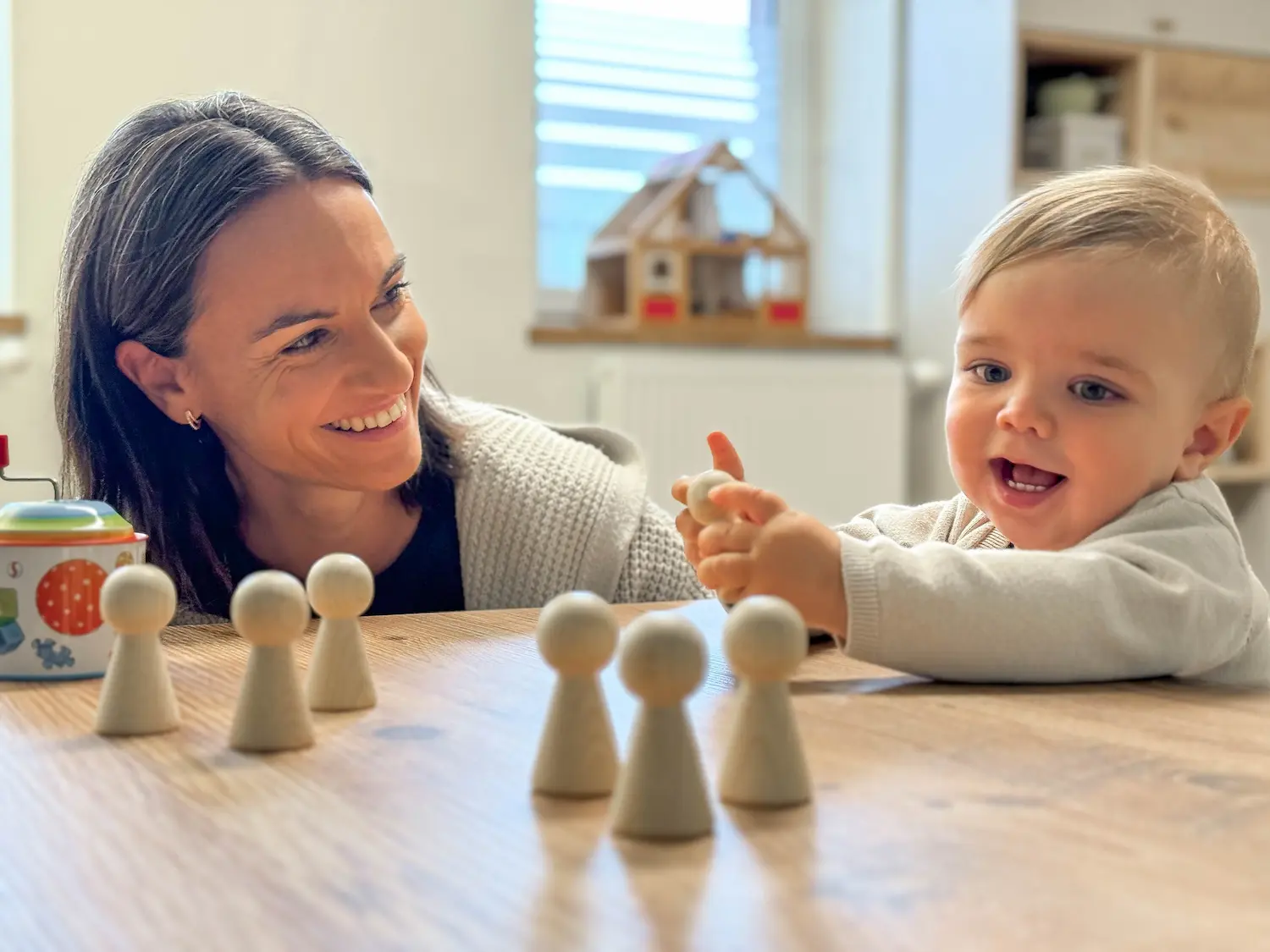 Dr. Martina Rinner lächelt ein Kleinkind an, das mit Holzfiguren spielt, und fördert dabei spielerisch die motorischen Fähigkeiten und das soziale Lernen in einer warmen Praxisatmosphäre.