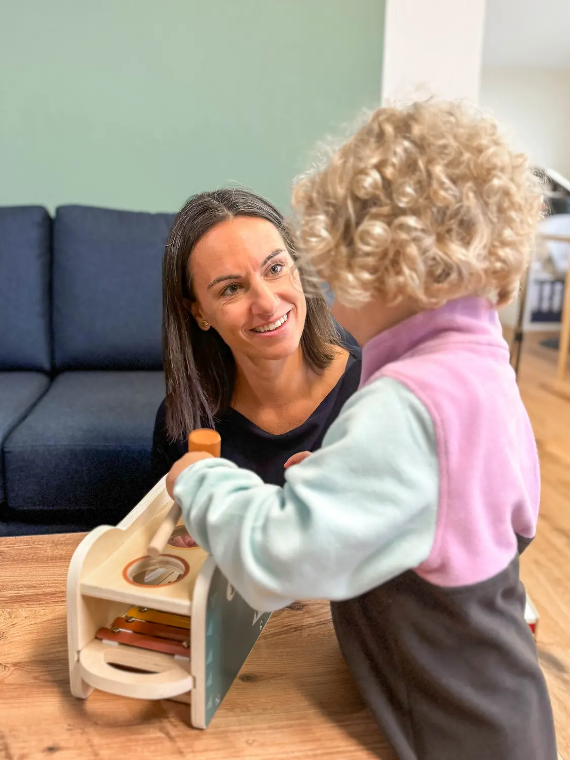 Dr. Martina Rinner beobachtet lächelnd ein Kind, das mit einem Holzspielzeug spielt, und fördert dabei die kindliche Neugier und Entwicklung in einer unterstützenden Praxisumgebung.