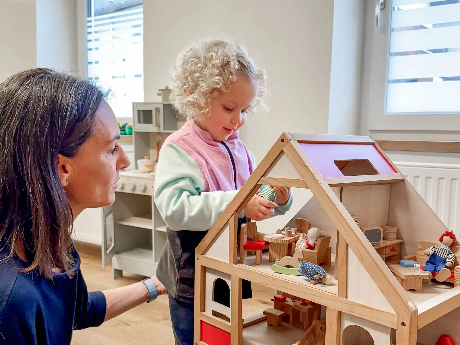 Dr. Martina Rinner spielt mit einem Kind, das ein Puppenhaus erkundet, und fördert dabei spielerisch die soziale Interaktion und das kreative Denken in einer sicheren Praxisumgebung.